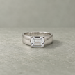 Silver ring with an emerald-cut clear solitaire stone and wide polished band.