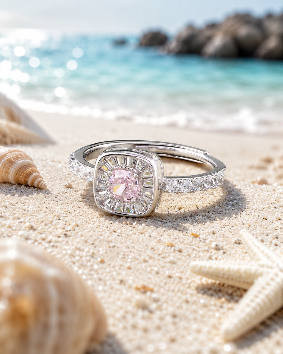 925 sterling silver halo ring with a pink cushion-cut center stone and sparkling cubic zirconia accents, displayed on sandy beach background with ocean waves.